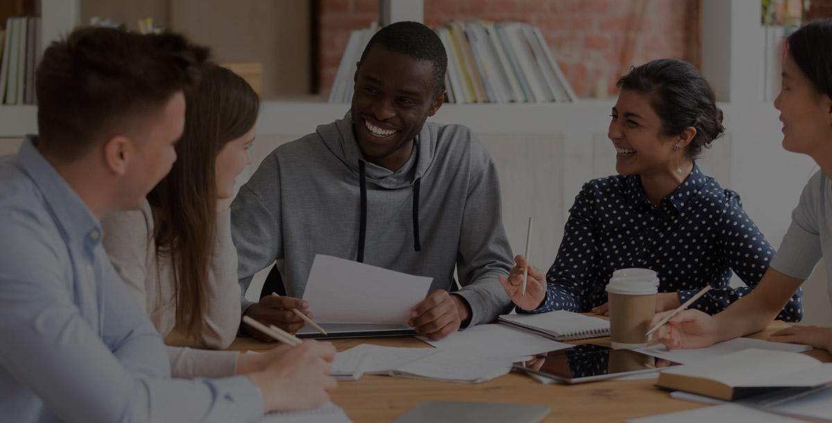 Five people sit at a table, engaged in a friendly discussion, with notebooks and papers in front of them.