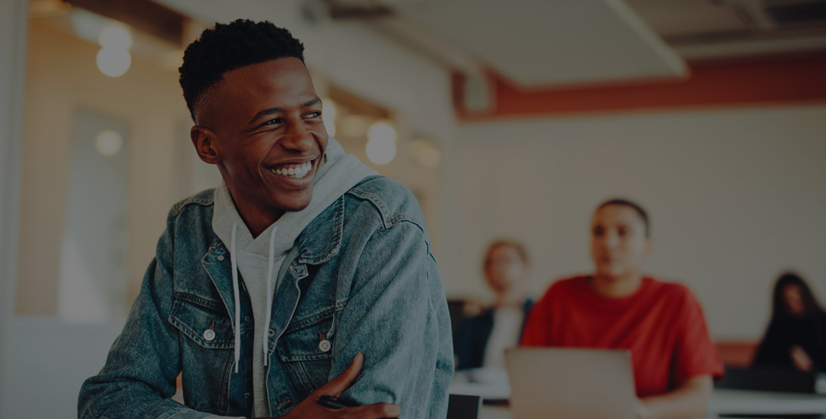 A smiling student in a denim jacket sits in a classroom, with others in the background.