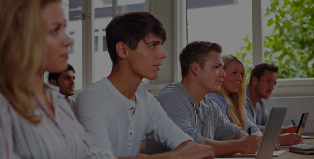 People sit in a classroom, attentively watching a presentation, with open laptops and notebooks in front of them.